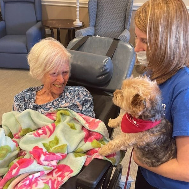 A woman holds a small brown dog up to a smiling elderly woman sitting in a wheelchair